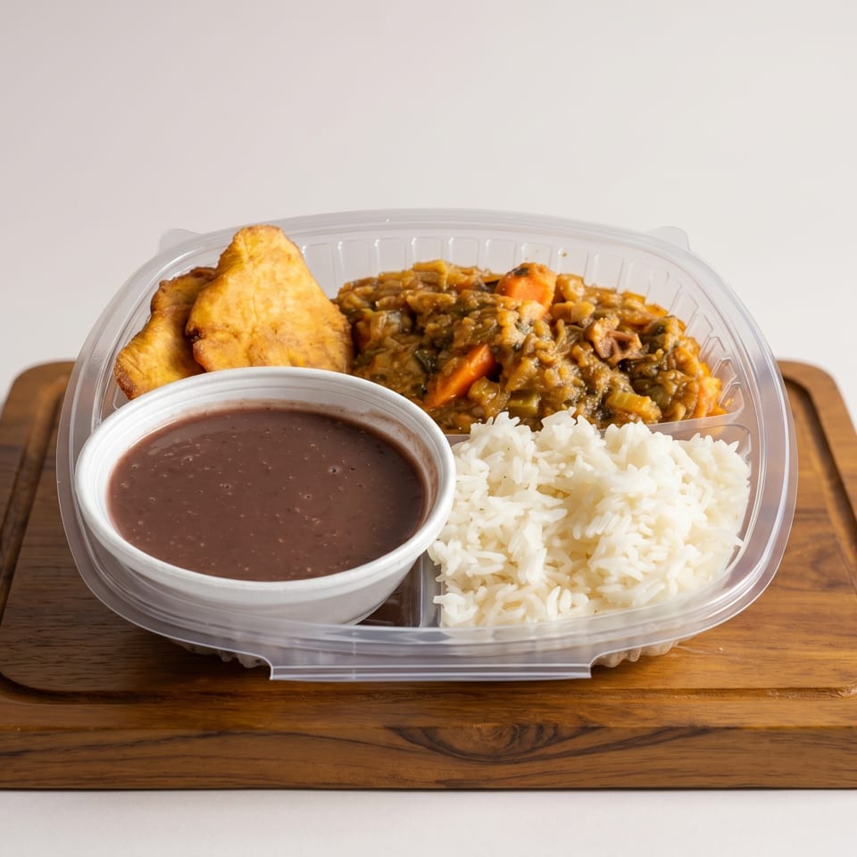 Takeout container with rice, stew, fried plantains, and a cup of black bean soup on a wooden board.