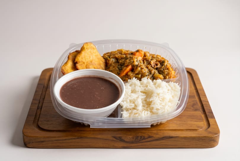 Takeout container with rice, stew, fried plantains, and a cup of black bean soup on a wooden board.