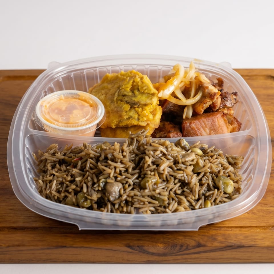 Takeout container with rice, fried plantains, chicken, onions, and a sauce cup on a wooden board.