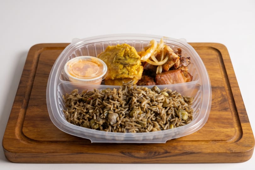 Takeout container with rice, fried plantains, chicken, onions, and a sauce cup on a wooden board.